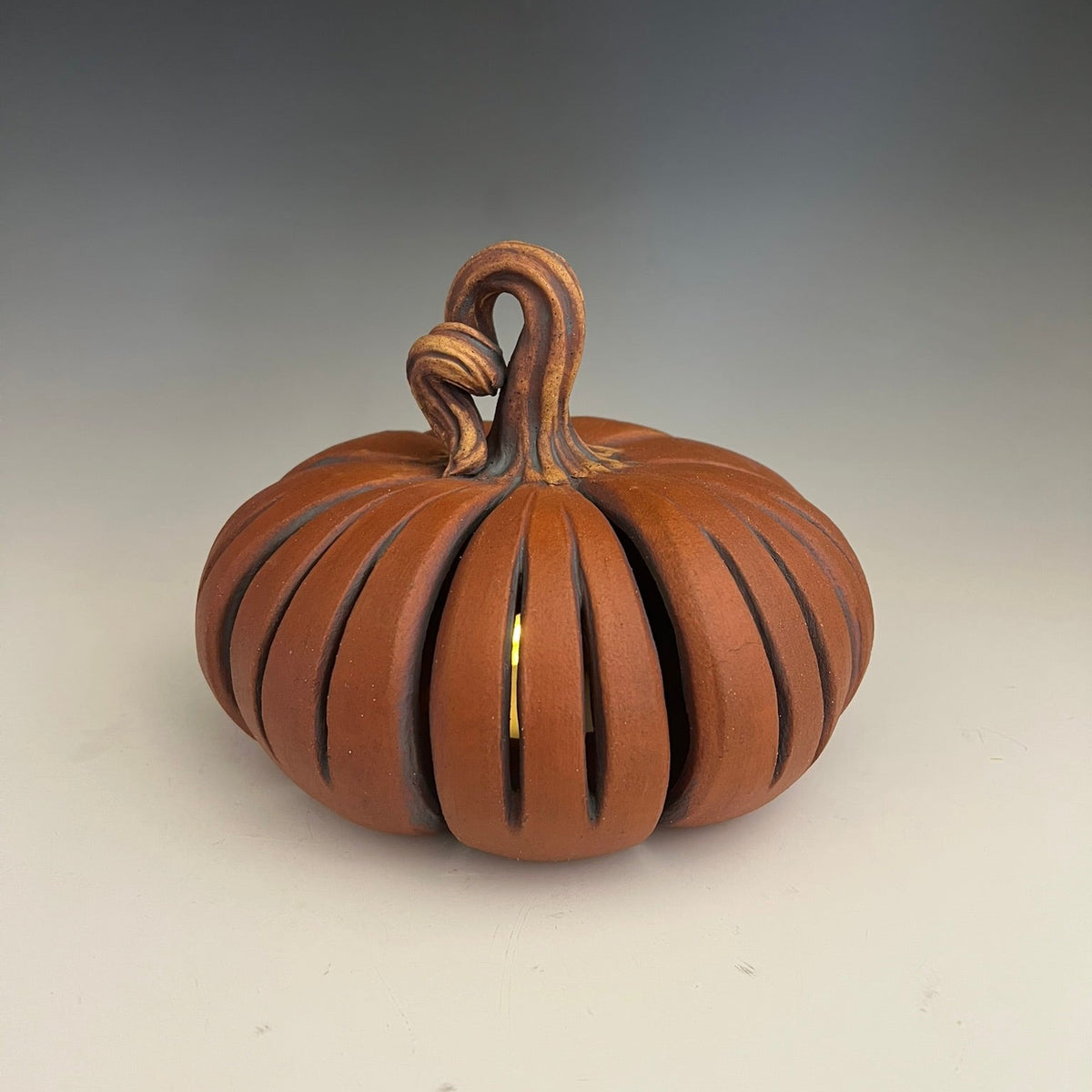 A carved pumpkin-shaped ceramic sculpture with vertical slits, lit from within, and set against a neutral gray gradient background.