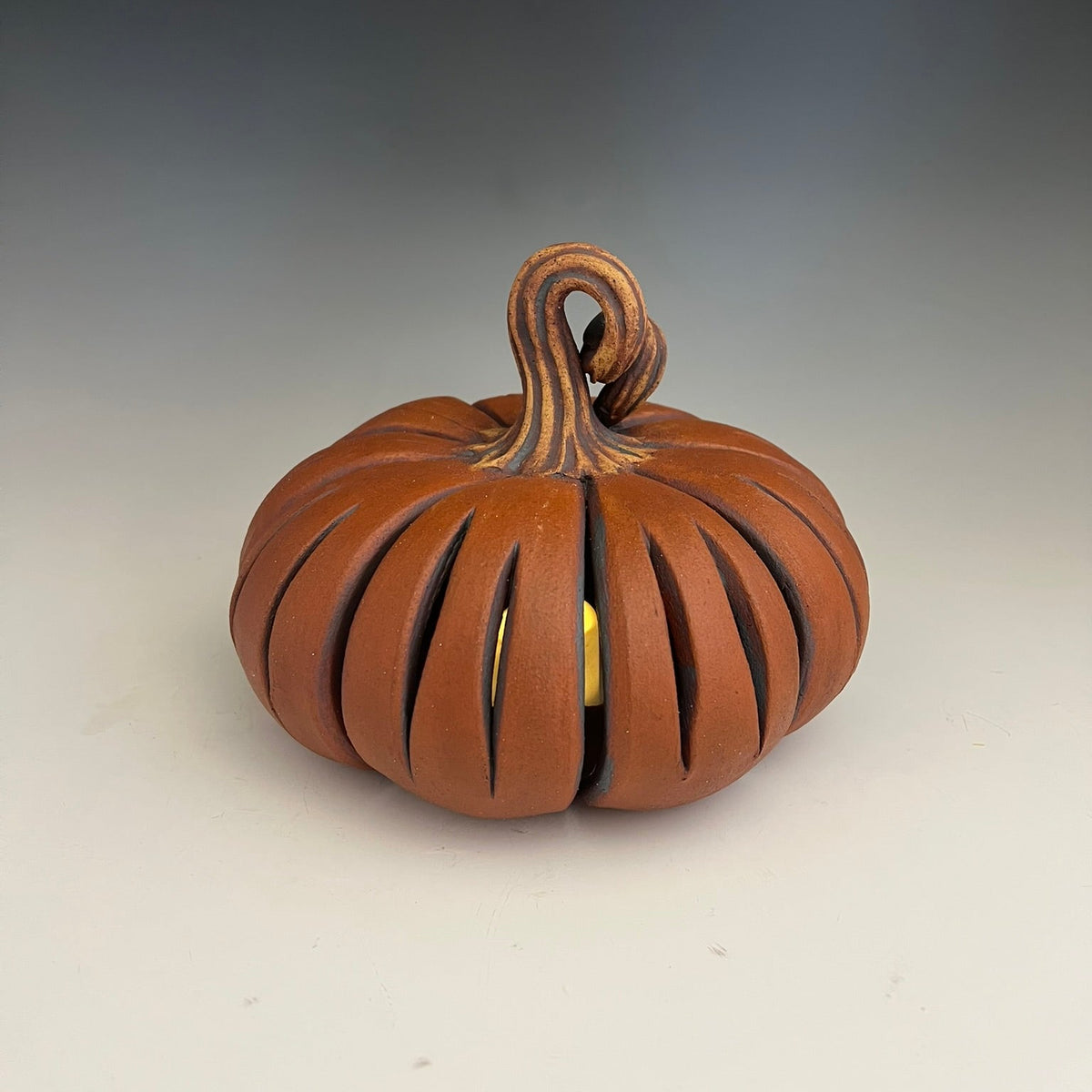 A decorative ceramic pumpkin figurine with a carved, ribbed body and a curled stem, shown on a neutral gray gradient background.