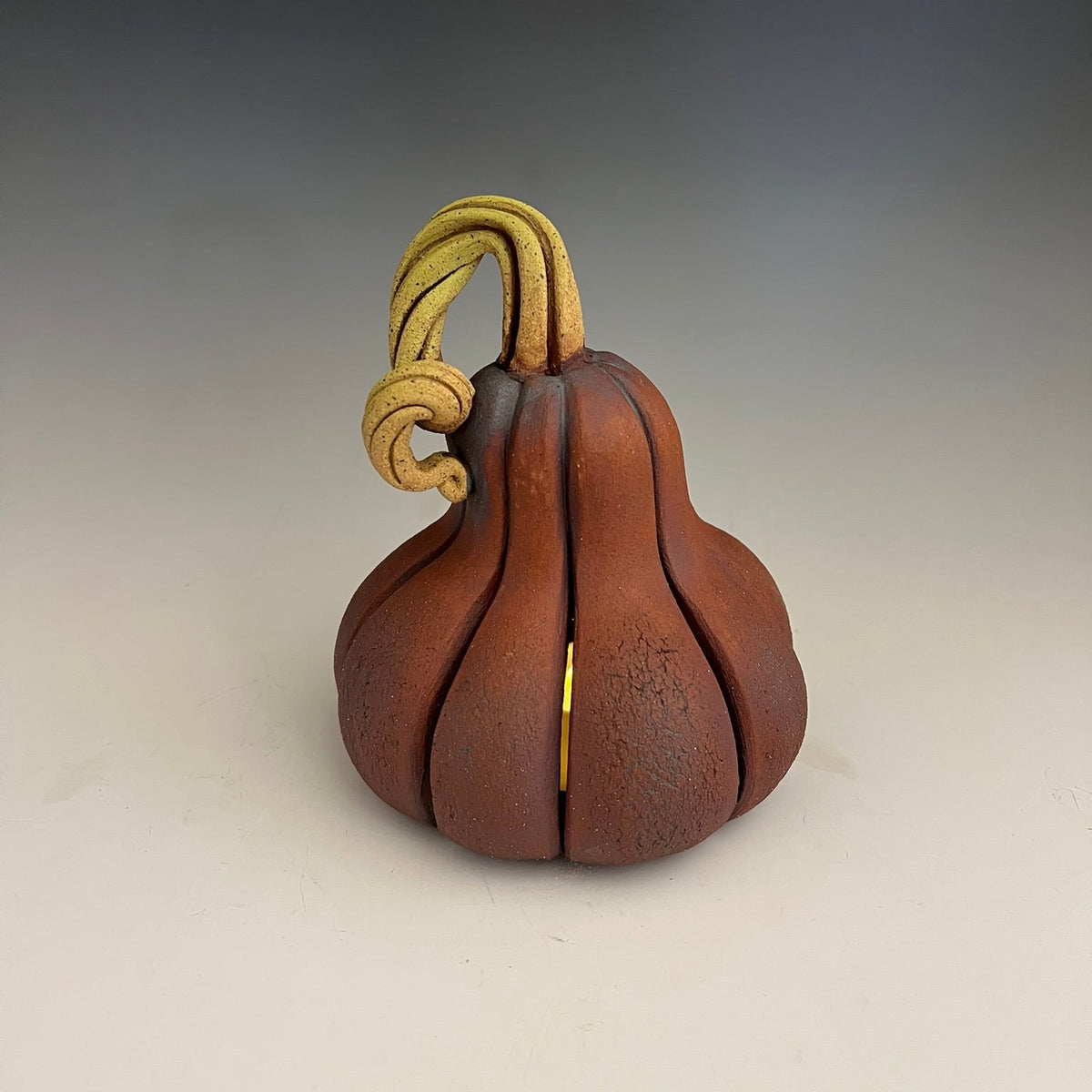 A small sculptural pumpkin-shaped object in warm reddish-brown with a twisted tan stem, photographed on a smooth gray-to-white studio background.