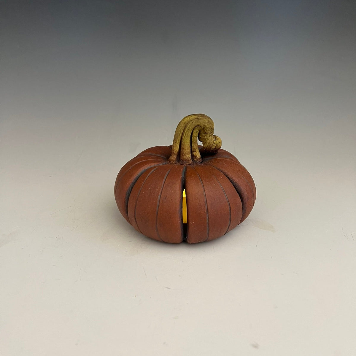 A small decorative pumpkin-shaped object with a ribbed brown-orange body, a tan stem, and a small gold accent visible on the front, photographed on a plain light background.