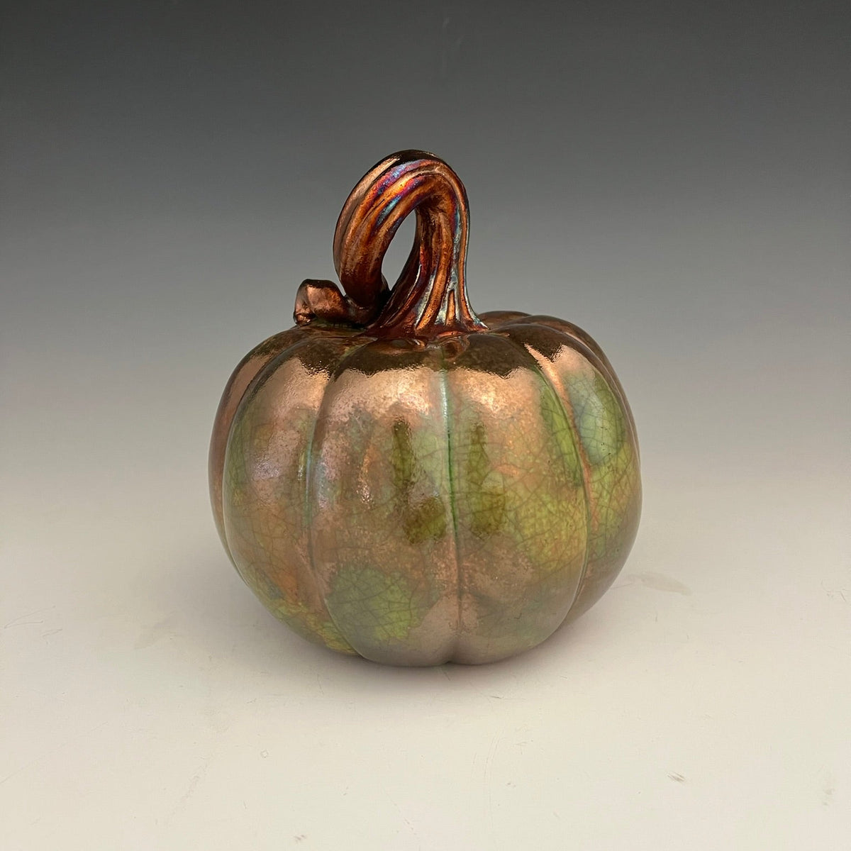 A decorative ceramic pumpkin with a glossy finish, featuring iridescent copper, bronze, and green leaf-like glaze patterns, set against a neutral gray studio background.