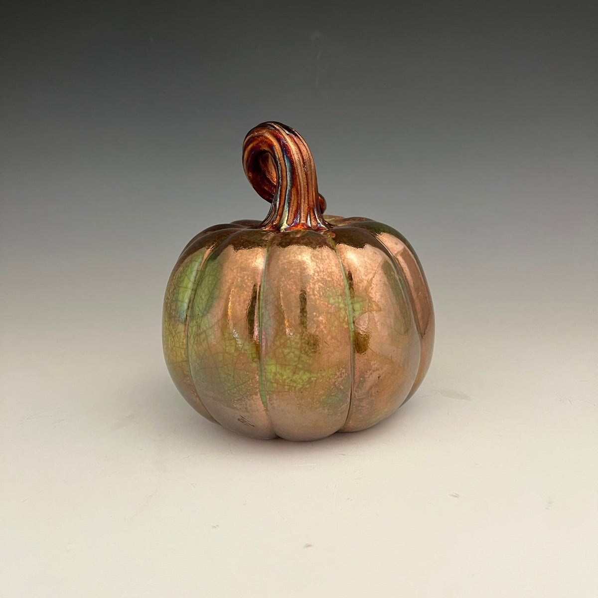 A decorative ceramic pumpkin with a metallic copper and green iridescent glaze, featuring a ribbed body and a curled handle, set against a neutral gray gradient background.