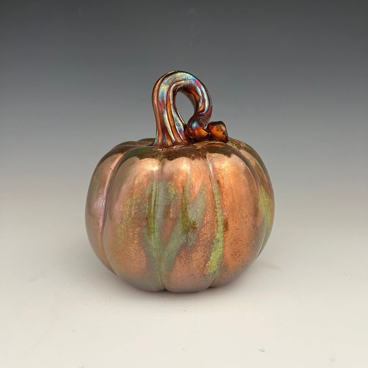 An iridescent copper-orange glass pumpkin figurine with a swirling green and purple sheen, featuring a looped stem, set against a neutral gray gradient background.