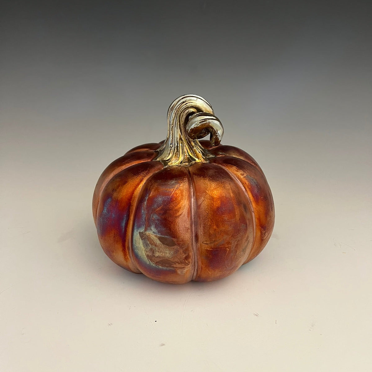 A decorative ceramic pumpkin with a glossy, iridescent finish showing orange, brown, and gold tones, set against a neutral gray gradient background.