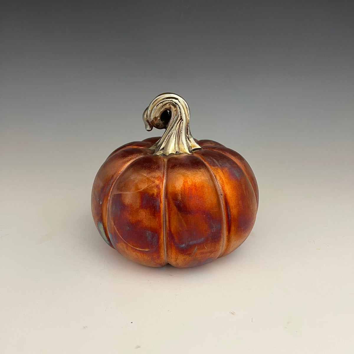 A decorative ceramic pumpkin figurine with a glossy, marbled orange and brown glaze and a metallic silver stem, set against a neutral gray gradient background.