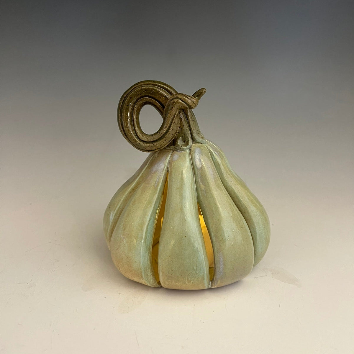 A ceramic decorative pumpkin sculpture with a glossy pale green and beige glaze, featuring a sculpted stem loop at the top and a warm yellow light glowing from within, set against a neutral gray gradient background.