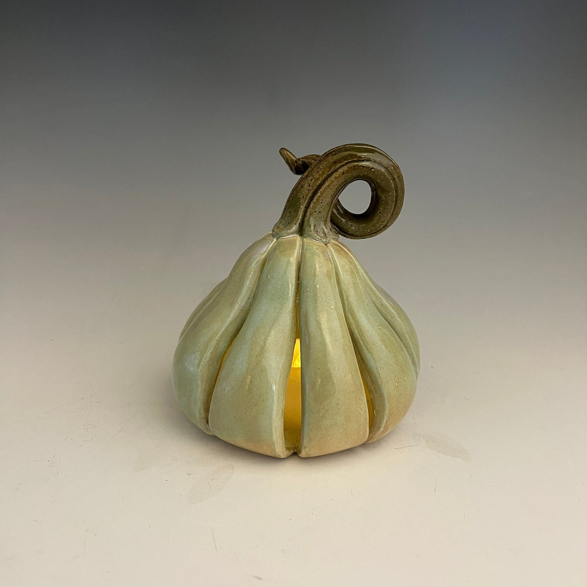 A small sculptural pumpkin-shaped vessel with a pale sage green glazed finish and a dark brown curled stem, shown on a neutral gray gradient background. A warm yellow light glows from within the center.