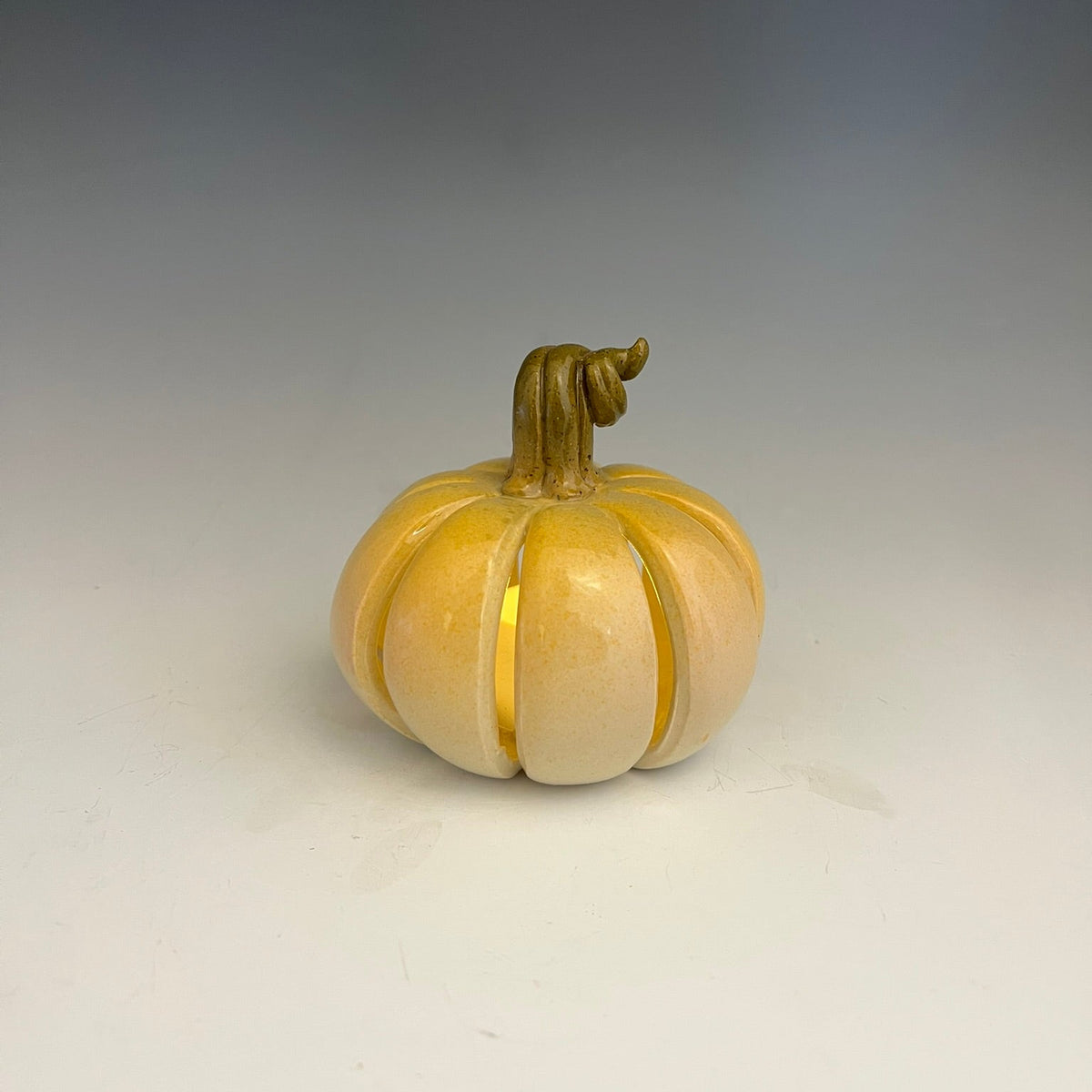 A small decorative pumpkin sculpture with a glossy finish, featuring a split design that reveals a warm yellow interior, set against a neutral gray gradient background.