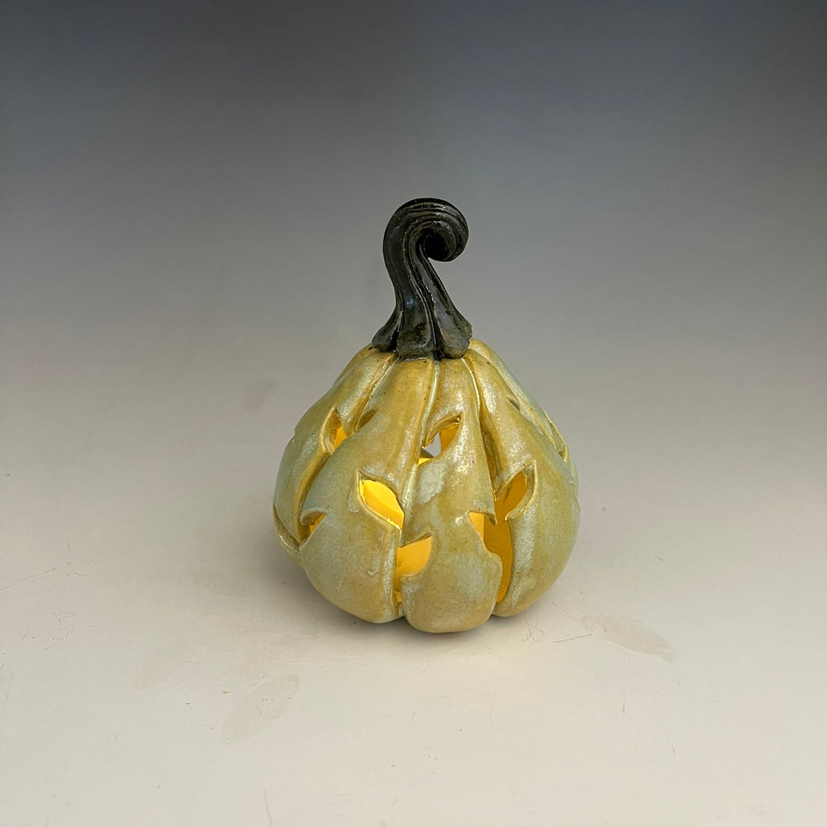 A small decorative ceramic pumpkin with a carved, cutout design that reveals a warm yellow glow from within, topped with a dark curled stem, set against a neutral gray background.