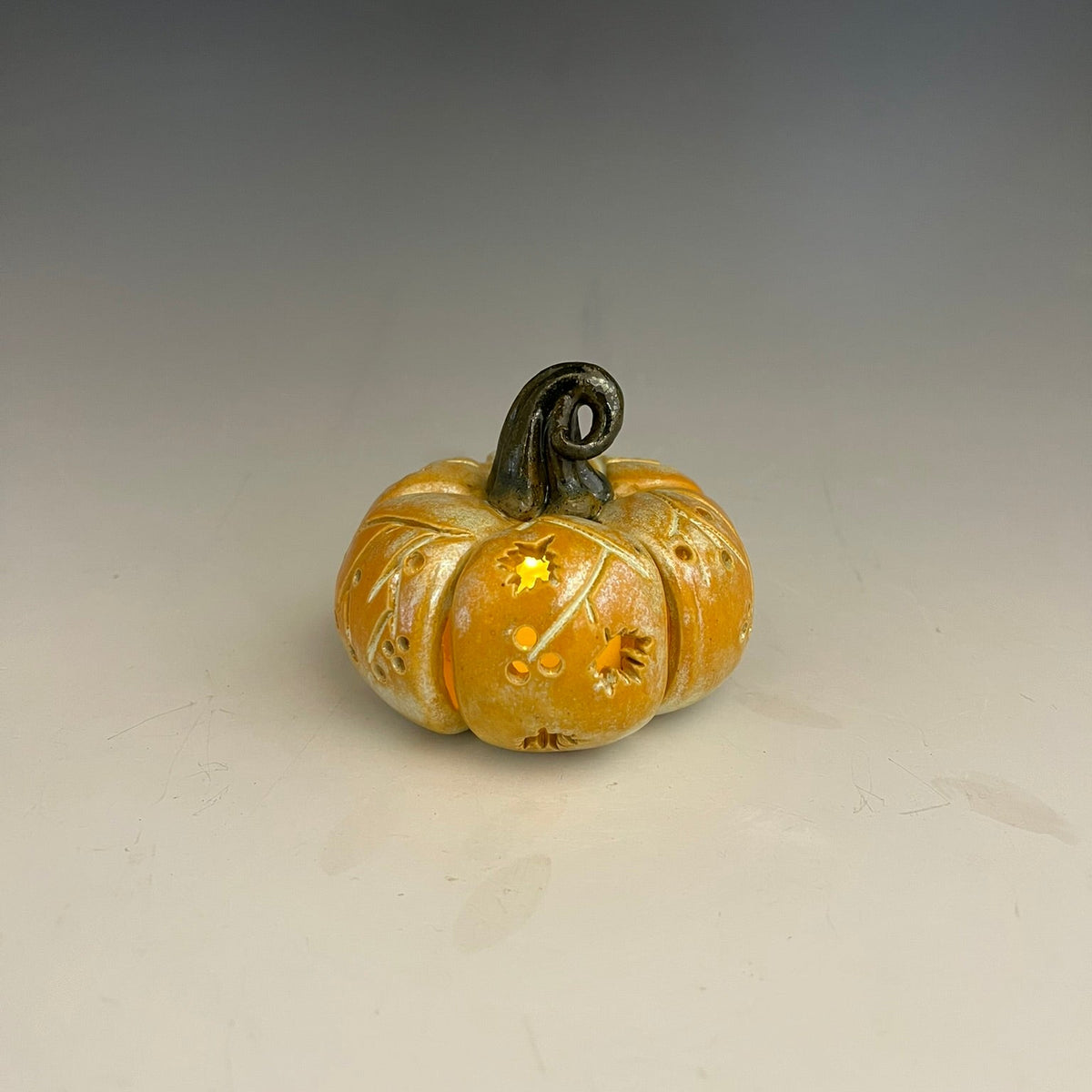 A small decorative ceramic pumpkin with a carved pattern and stem, lit from within with a warm yellow glow, set against a neutral gray gradient background.