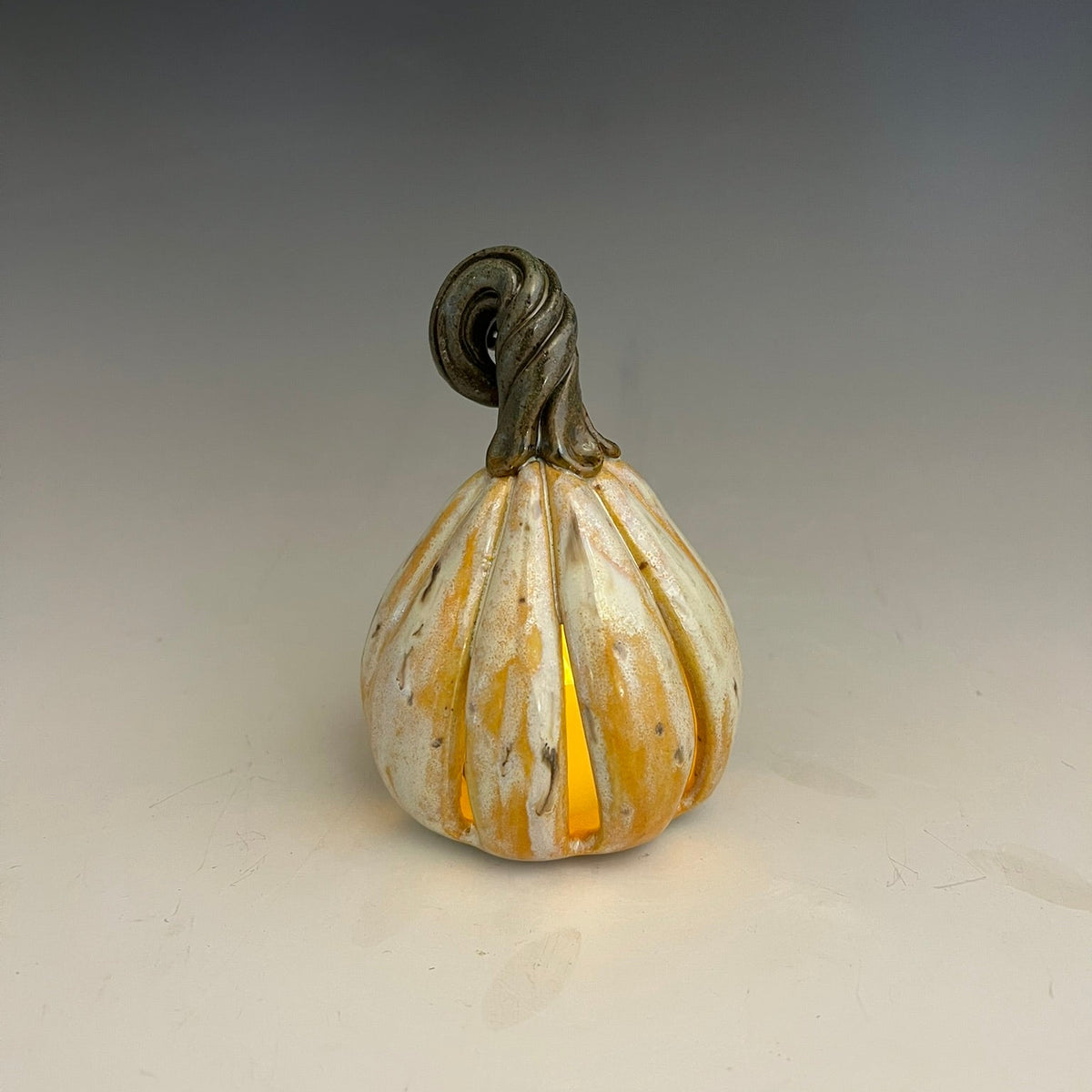 A small decorative ceramic pumpkin with a ribbed body, a dark gray twisted stem, and a warm yellow-orange glow emanating from within, set against a neutral gray gradient background.
