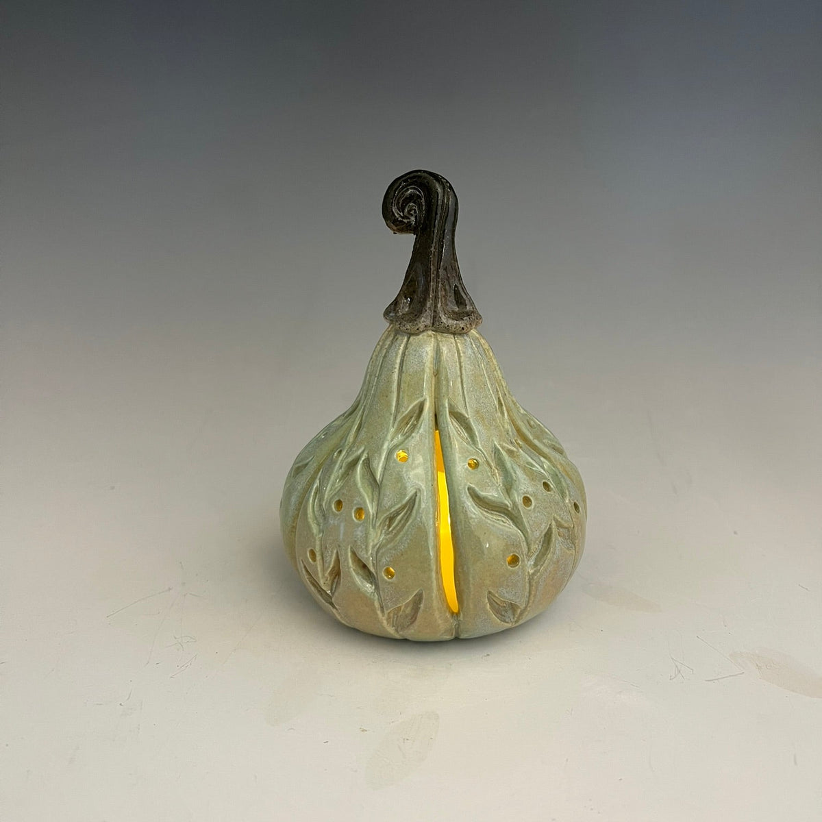 A ceramic decorative gourd figurine with a pale sage green body, textured leaf patterns, a dark brown twisted stem, and a bright yellow vertical accent running down the center.