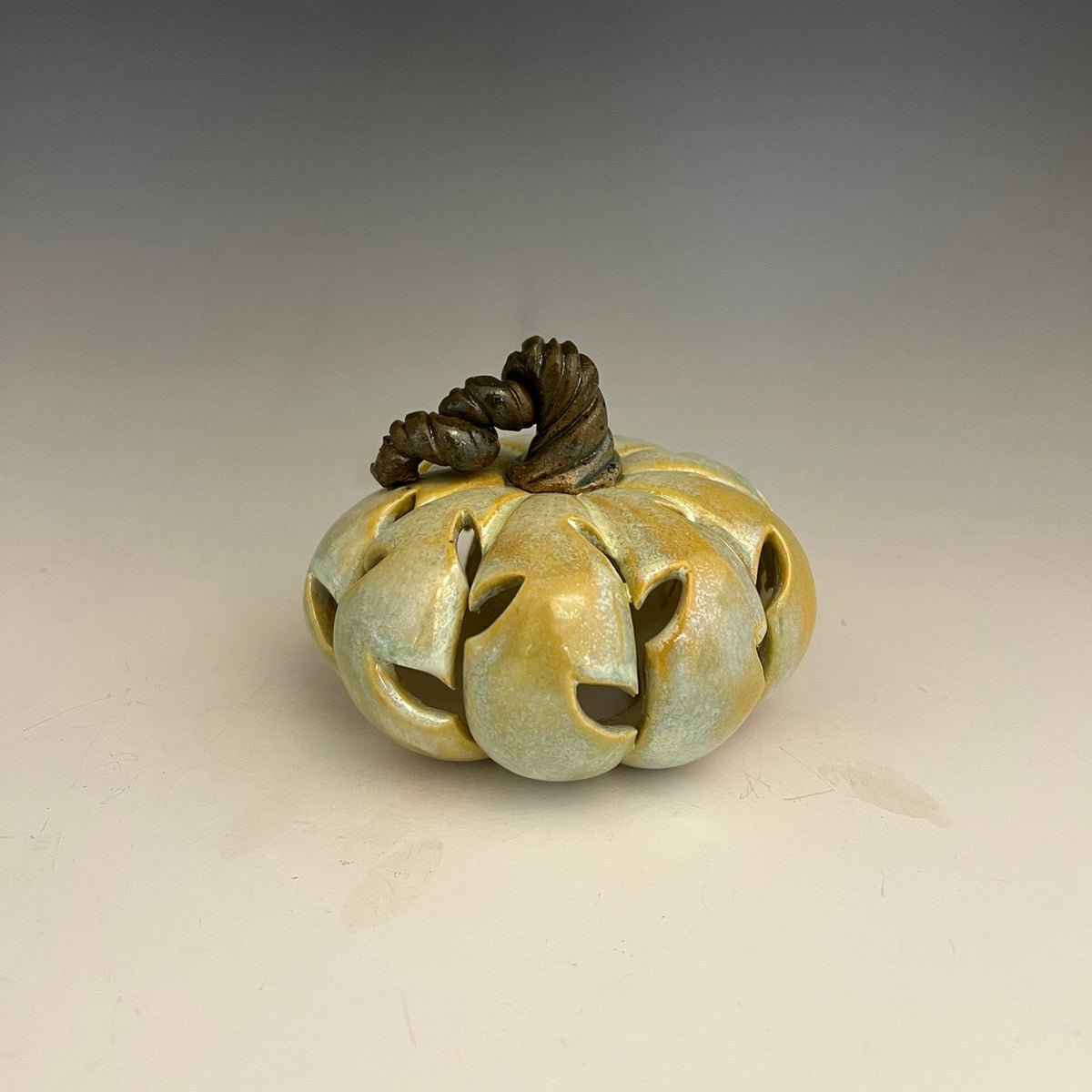 A decorative ceramic pumpkin sculpture with carved leaf design, featuring a glossy pale green and gold glaze with a dark brown stem, set against a neutral gray gradient background.