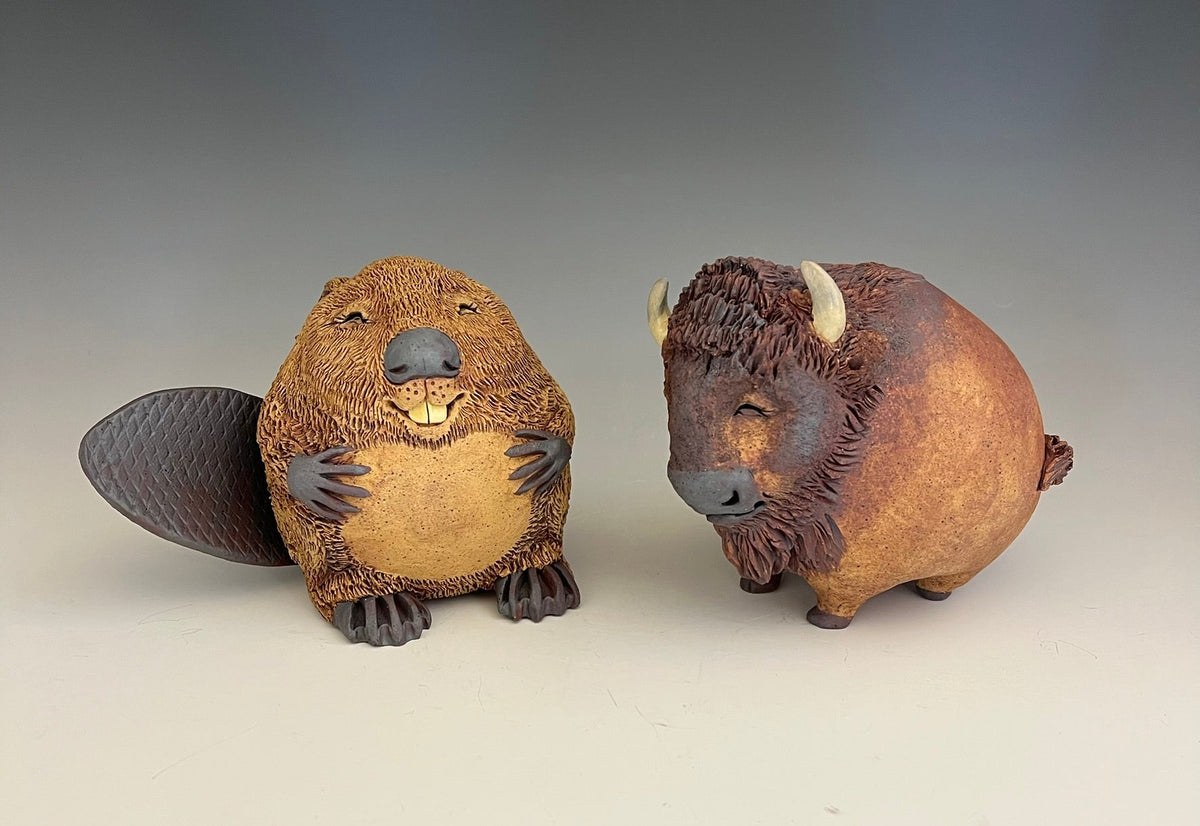 Two small animal figurines, a brown beaver with a dark tail flap on the left and a reddish-brown bison with white horns on the right, displayed against a neutral gray studio background.