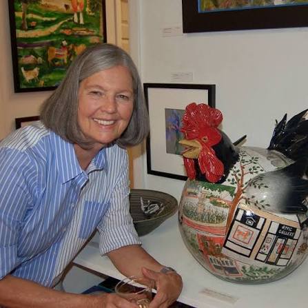 Terri Axness smiling at a table next to a large decorative ceramic rooster figurine. The scene is inside an art gallery with framed paintings on the wall in the background.