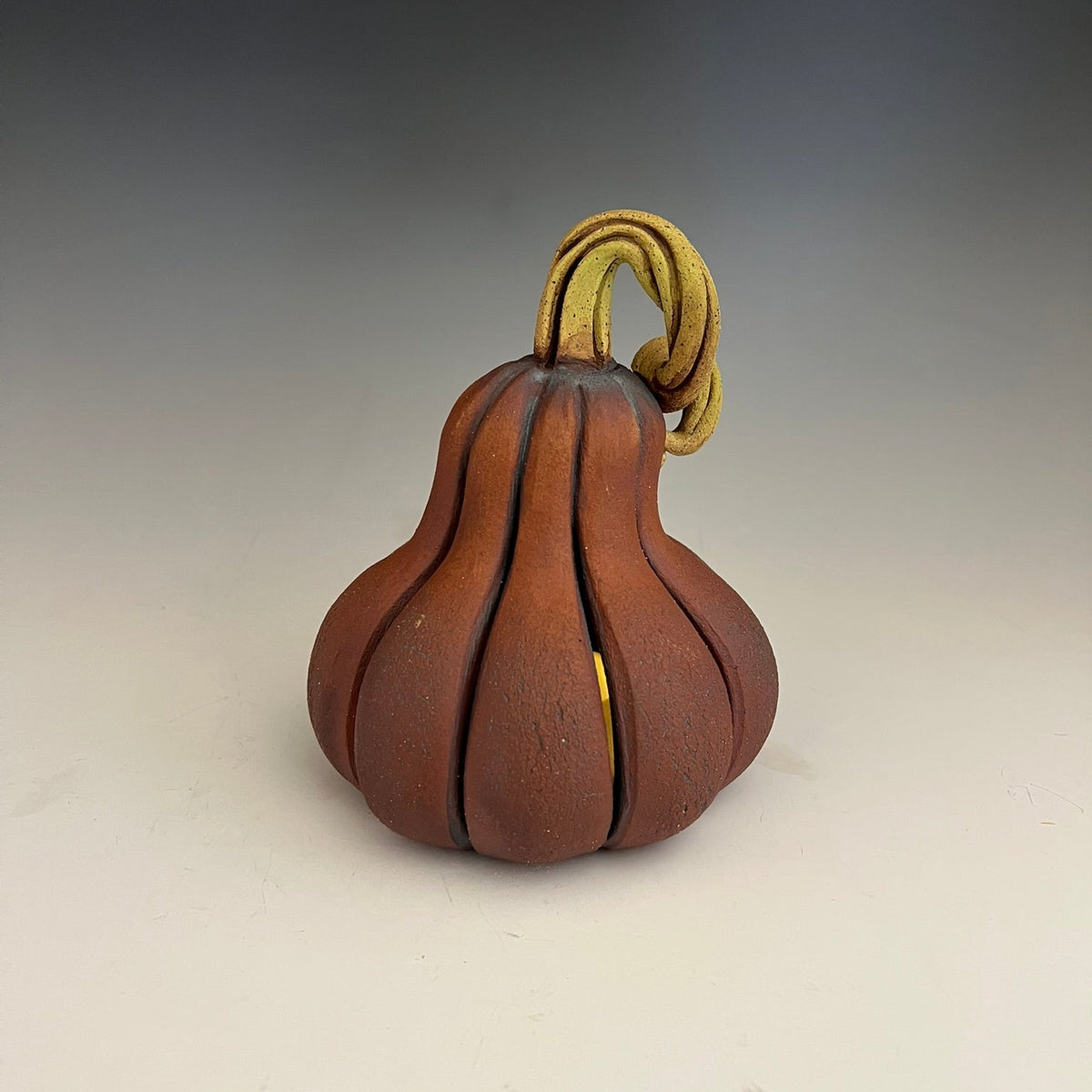 A small sculptural pumpkin-shaped object with a red-brown body, a tan looped stem, and subtle gold-toned accents, photographed against a dark-to-light gradient studio background.
