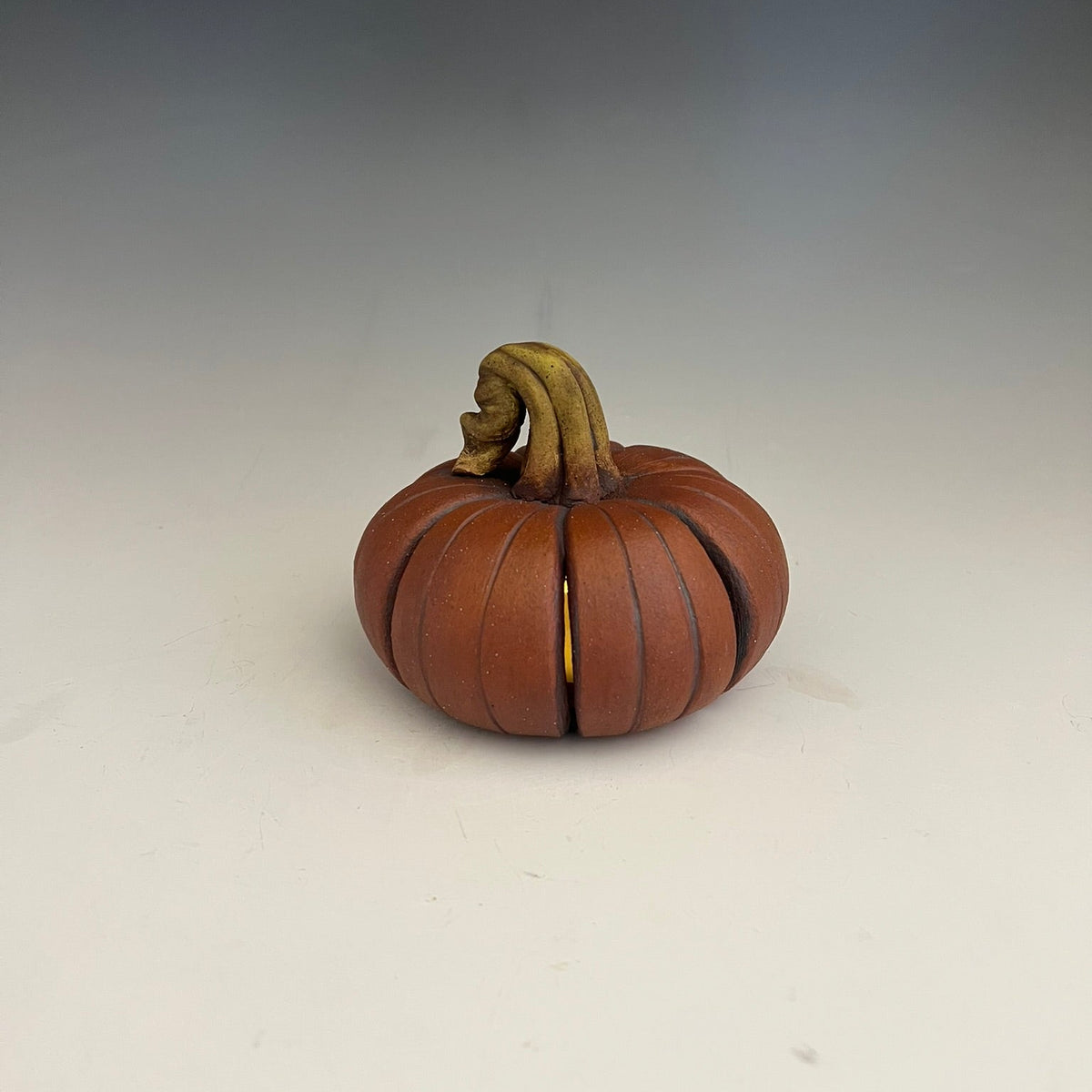 A small decorative pumpkin-shaped object with a ribbed orange-brown body and a curved tan stem, photographed on a plain light background with a dark gradient at the top.