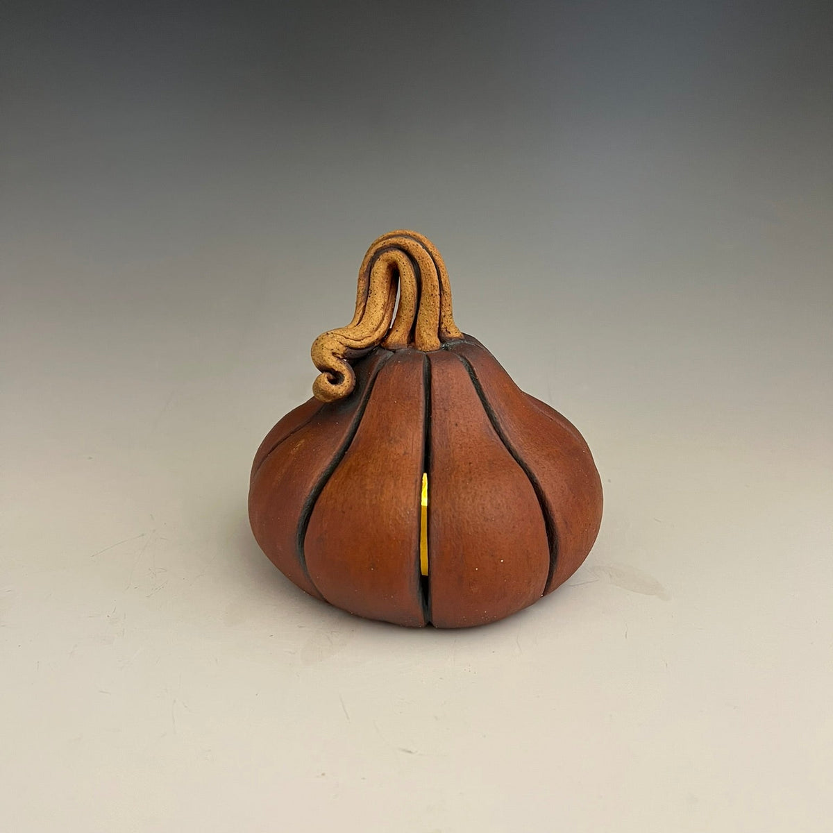 A small decorative reddish-brown pumpkin-shaped object with a curved tan stem, shown on a neutral gray studio background.