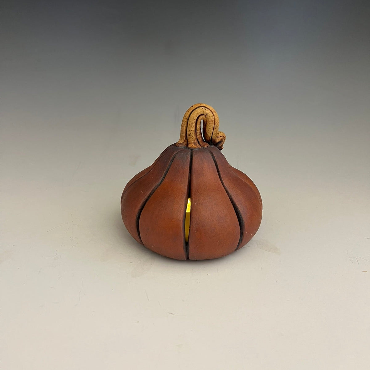 A small decorative pumpkin-shaped object with a warm brown body, dark seam lines, and a curled tan stem, photographed on a smooth light gray to white studio background.