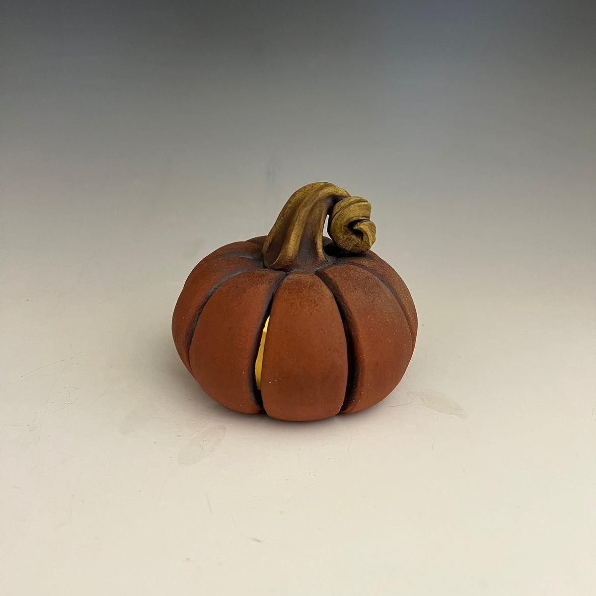 A small sculpted pumpkin figurine in terracotta brown with a tan stem, photographed on a neutral light gray to white studio background.