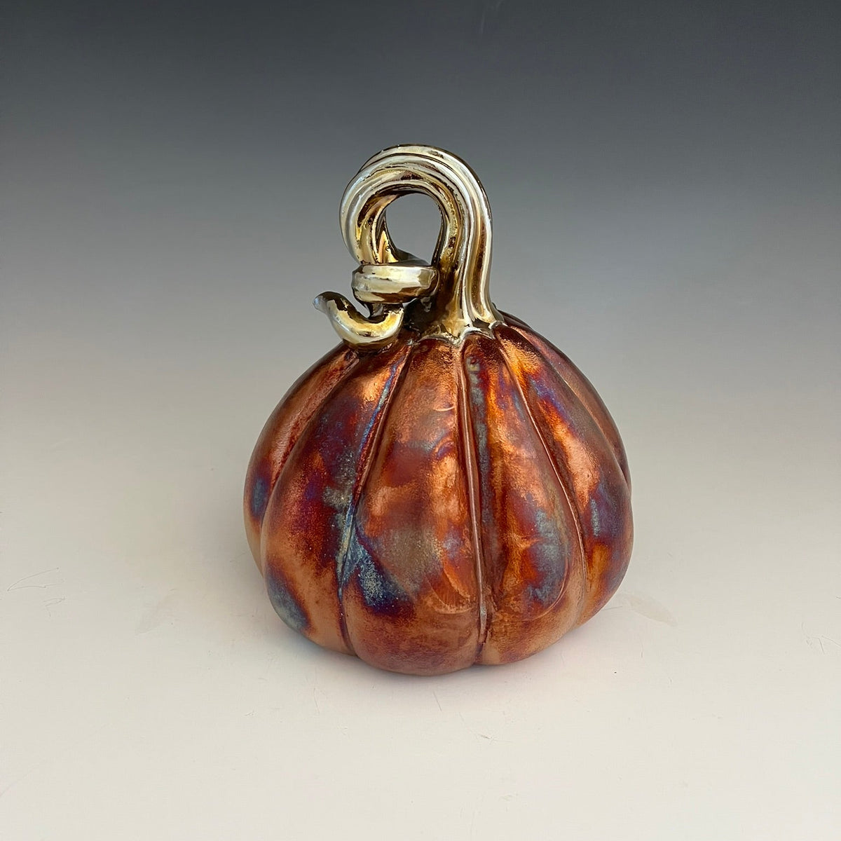 A decorative ceramic pumpkin figurine with a metallic copper and amber glaze, featuring a glossy silver handle, set against a neutral gray gradient background.