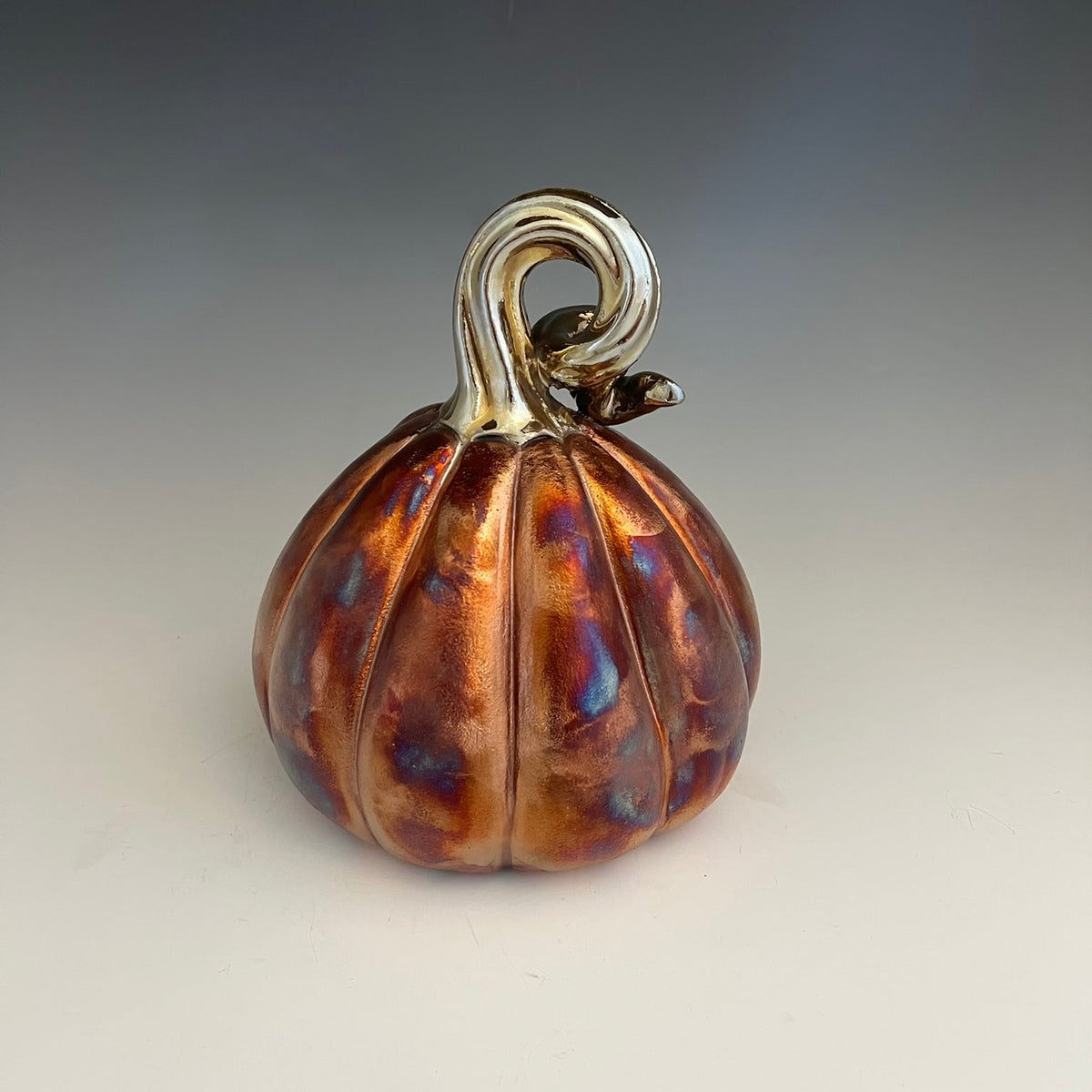 A small decorative pumpkin figurine with a metallic copper and amber body, iridescent purple-blue highlights, and a silver-toned stem, photographed on a neutral gray-to-white studio background.