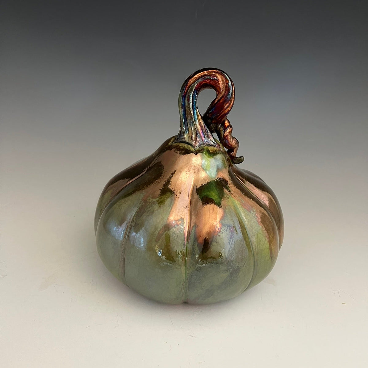 An iridescent ceramic pumpkin sculpture with a glossy finish, featuring shifting colors of green, bronze, and copper, set against a neutral gray gradient background.