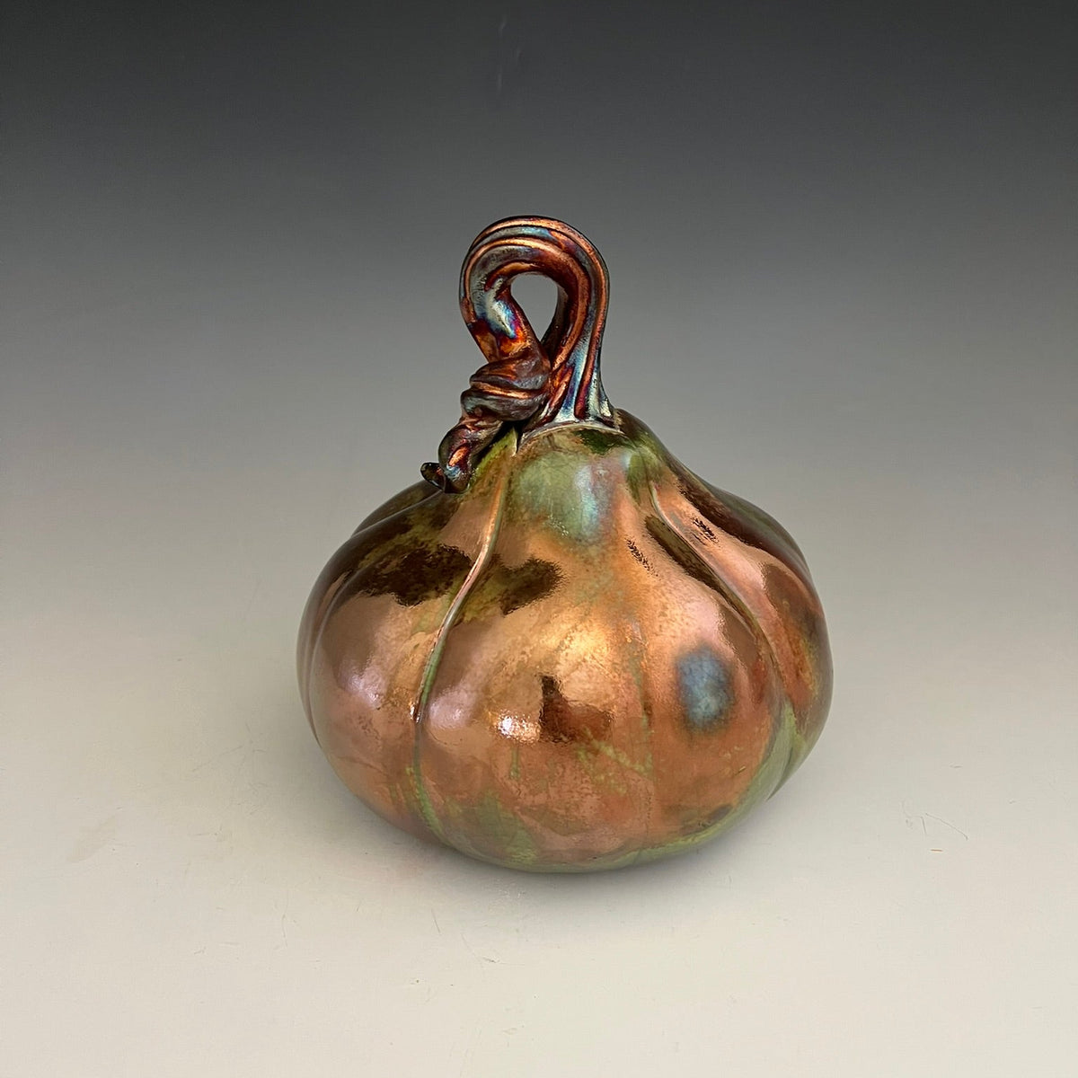 A decorative sculptural pumpkin with a glossy copper and green iridescent glaze, featuring a curled copper stem, set against a neutral gray gradient background.
