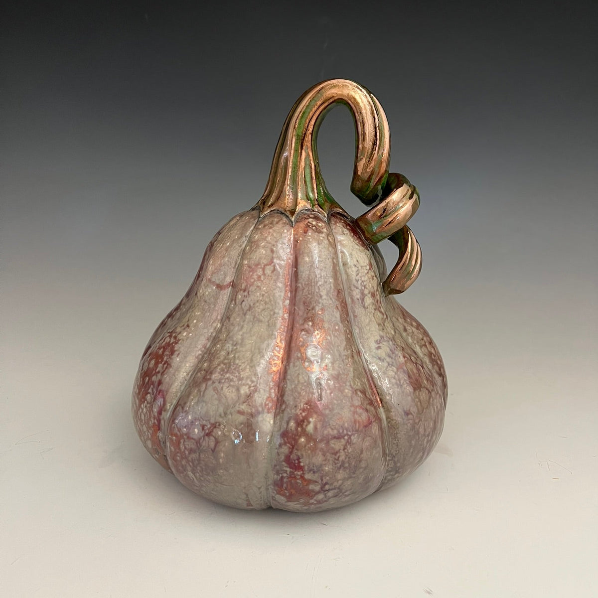 A decorative ceramic pumpkin figurine with a glossy finish, featuring a mottled gray and pink body with copper-toned ridges and a curled gold stem, set against a dark-to-light gray gradient background.