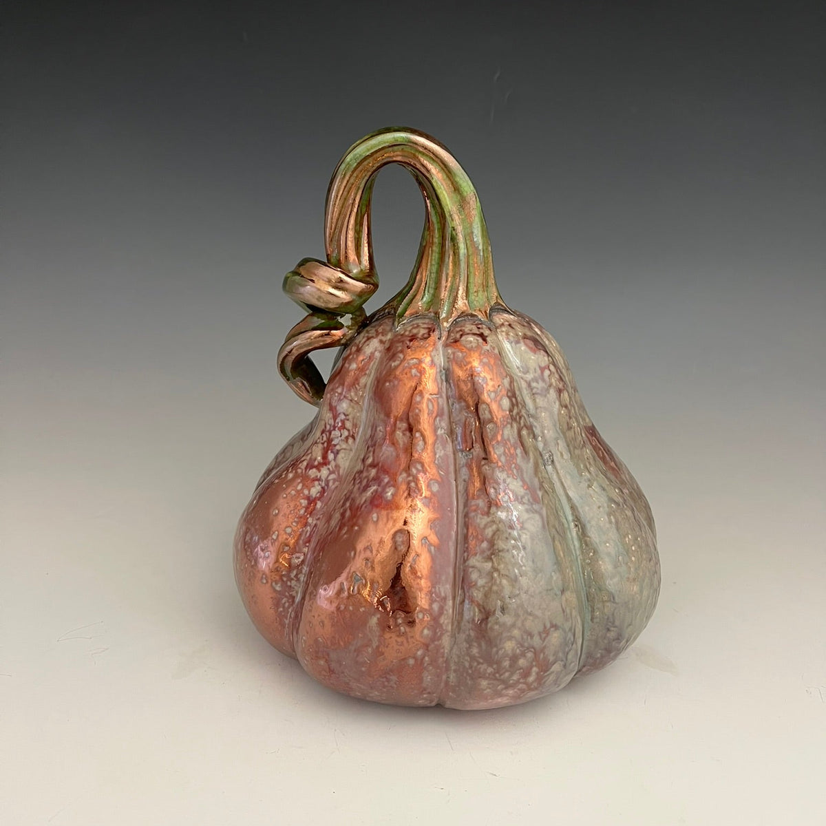 A decorative ceramic pumpkin figurine with a glossy, metallic finish, featuring a copper-orange body with green and gold accents on the stem and ridges, set against a dark-to-light gray gradient background.