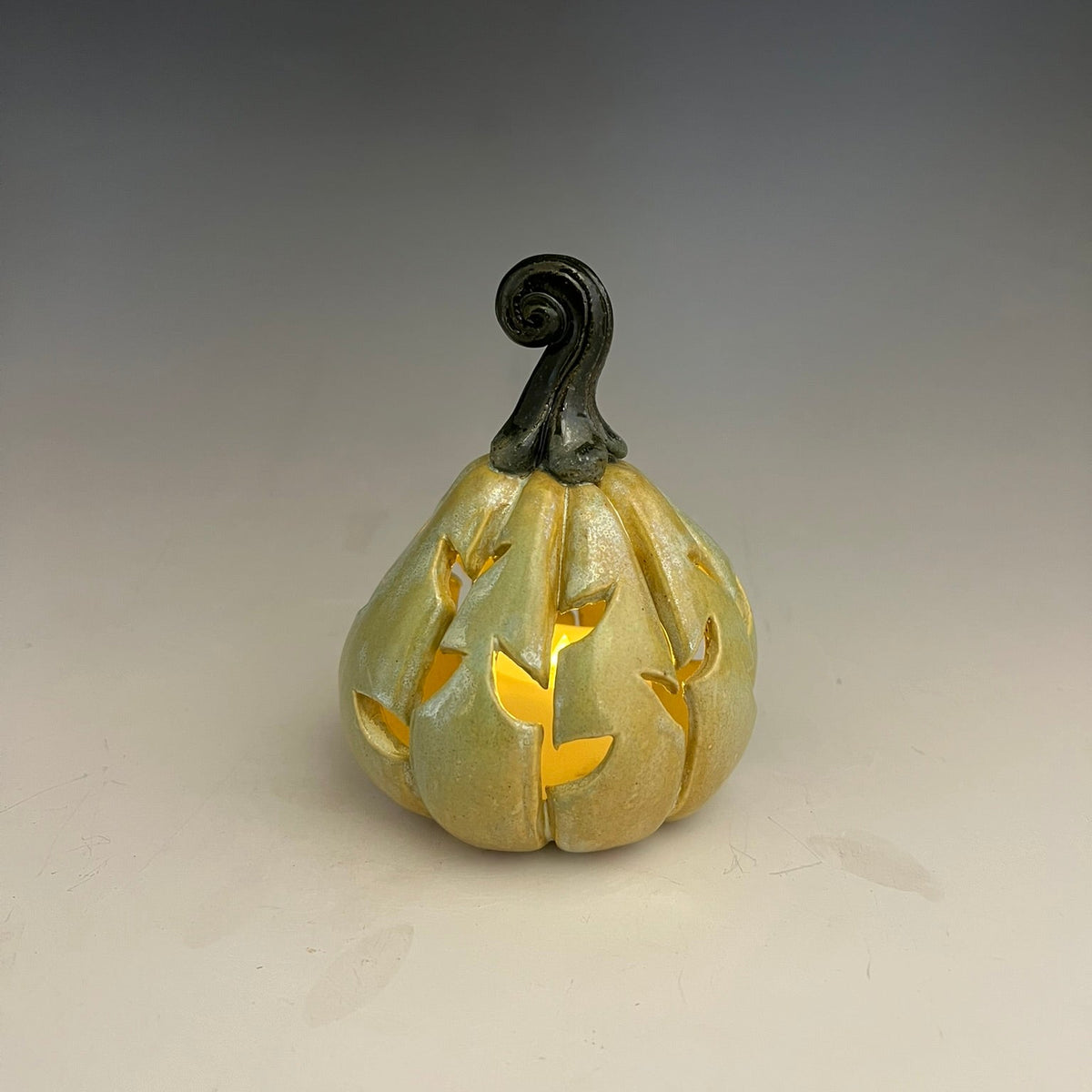 A small decorative ceramic pumpkin with a carved leaf pattern, lit from within to glow with a warm yellow light, set against a neutral gray gradient background.