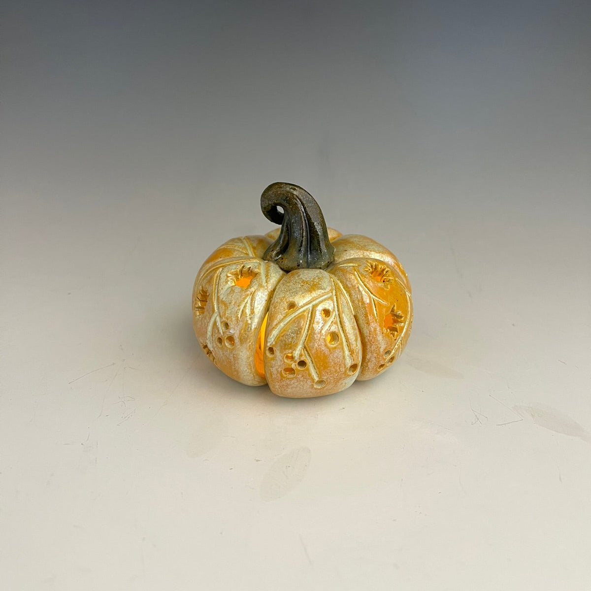 A small decorative carved pumpkin figurine with a dark stem, featuring cutout details that reveal warm orange light inside, set against a neutral gray gradient background.