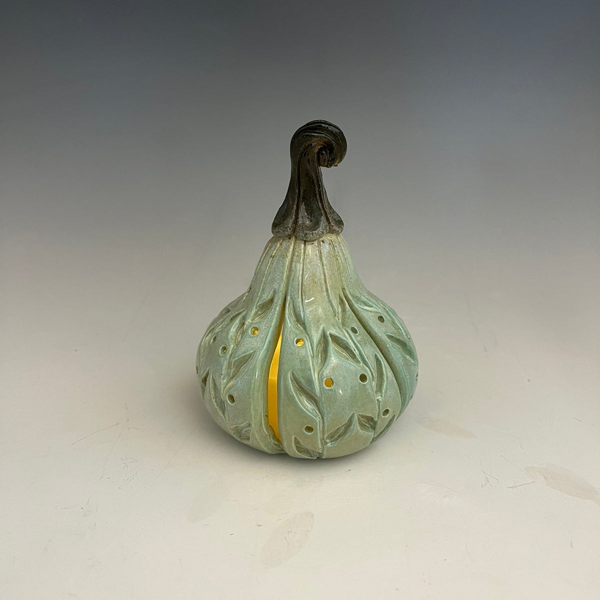 A ceramic decorative gourd figurine with a pale mint green glazed body, leaf-like relief patterns, a dark brown twisted stem, and a vertical gold accent stripe, set against a neutral gray gradient background.