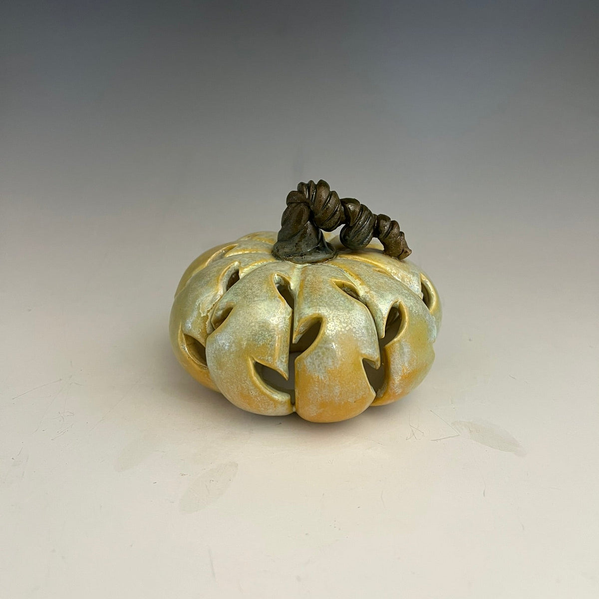 A carved ceramic pumpkin figurine with a textured, metallic gold and olive-green glaze, featuring a dark brown stem, set against a neutral gray gradient background.